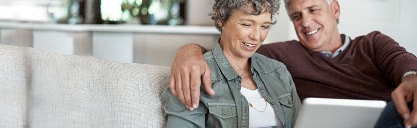 Mature couple sit on their couch looking at their computer while man has his arm around the woman.