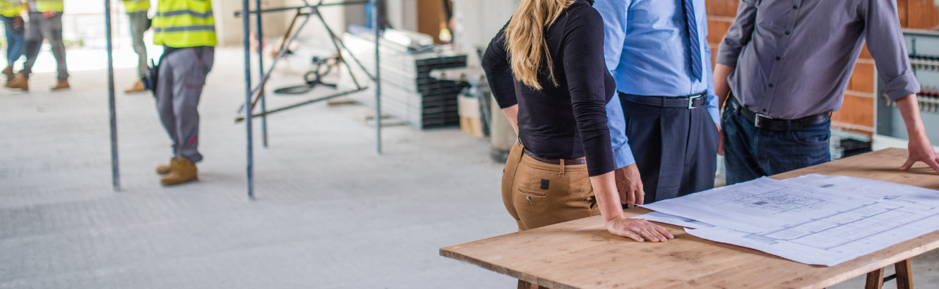 Three individuals stand by blueprints discussing what they see. While construction is taking place in the background.