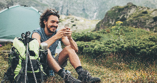 A man on his phone while he sits in the wilderness next to his backpack and tent.